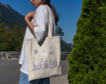 May include: Beige tote bag featuring a lavender field and butterfly design. The bag has a rectangular shape with a wide opening and long straps. The design includes purple lavender flowers and butterflies. The bag is carried by a person.