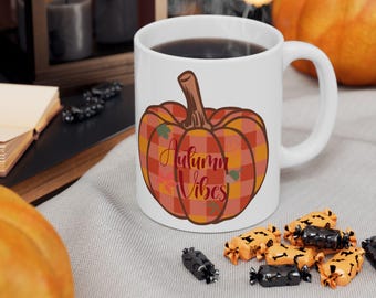 Può includere: Tazza in ceramica bianca con manico, riempita di liquido scuro. La tazza presenta un disegno a quadri di zucca con le parole "Autumn Vibes" in corsivo. Caramelle e una zucca sullo sfondo.