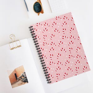 Könnte beinhalten: Ein pinkfarbenes Notizbuch mit Spiralbindung und einem Muster aus rot-weißen Zuckerstangen und kleinen weißen Punkten. Das Notizbuch ist geöffnet und zeigt leere weiße Seiten. Eine goldfarbene Sonnenbrille und eine Büroklammer sind ebenfalls zu sehen.