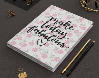 May include: A floral-patterned journal with the words "Make Today Fabulous" in black script. The journal features pink flowers and green leaves on a white background. Gold and black pencils and gold binder clips are also visible.