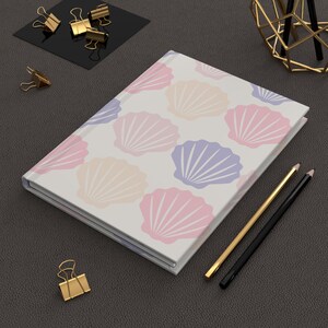 May include: A journal with a white cover featuring a pattern of pastel-colored seashells in pink, peach, and purple. Gold and black pencils and gold binder clips are arranged around the journal on a dark gray surface.