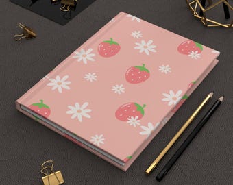 May include: A closed pink journal with a pattern of strawberries and white daisies. Two pencils, one gold and one black, are next to the journal. Gold binder clips are scattered around the journal.