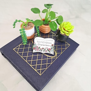 May include: A book-shaped enamel pin with the text "Emotionally attached to fictional characters" and pink flowers. The pin is on a blue book with gold accents and surrounded by four small potted plants.