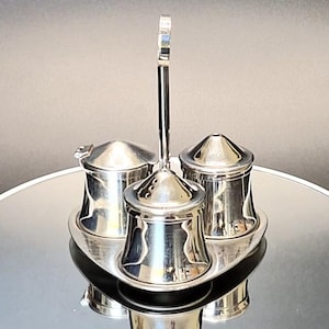 May include: A silver-coloured condiment set featuring three small containers arranged on a triangular tray. The set includes a central shaker with a perforated lid and a tall handle, flanked by two lidded pots with smooth, polished metallic surfaces.