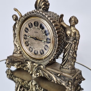 May include: A metallic gold-toned mantel clock featuring a circular face with black Roman numerals and the brand name Coral. The clock is flanked by two standing figures and sits on an ornate base with intricate scrollwork and a textured finish.