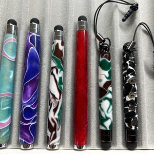 Puede incluir: Una colección de coloridos lápices ópticos con puntas plateadas. Los lápices presentan varios diseños, incluyendo remolinos de azul, morado, verde y blanco, así como un lápiz rojo liso. Algunos lápices tienen un cable negro con un conector para auriculares.