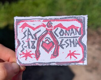 May include: A hand-held, rectangular art print with a red and black design on white paper. The print features stylized text and a central figure with a heart shape. The text includes the words "SA 18" and "CONAN KSHA".