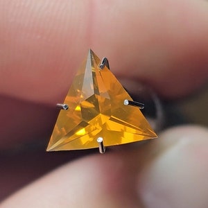 May include: A triangular, faceted gemstone with a golden orange color. The gemstone is set in a prong setting with four prongs.
