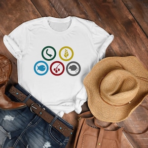 Könnte beinhalten: Weißes T-Shirt mit fünf bunten Kreisen, die jeweils ein anderes Symbol enthalten: einen Fisch, einen Wurm, eine Beerenkette, eine Weizenähre und eine Maus. Das Shirt liegt auf einer Holzoberfläche mit einer braunen Ledertasche, einer blauen Jeans und einem braunen Strohhut.