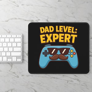 May include: Black rectangular mousepad with the text "DAD LEVEL: EXPERT" in yellow. A blue controller with sunglasses and a mustache is below the text. A white keyboard is visible on the left.
