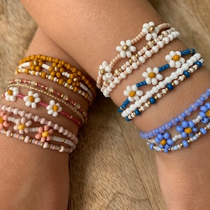 May include: A set of colorful beaded bracelets with daisy flower accents. The bracelets are made with small beads in shades of pink, blue, yellow, and white. The daisy flowers have white petals and yellow centers.