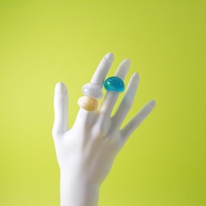 May include: A hand model wearing three rings. The rings are stacked on the middle finger. The top ring is blue, the middle ring is gold, and the bottom ring is silver.