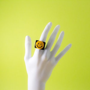 May include: A black ring with a yellow smiley face on it. The ring is worn on the index finger of a hand.