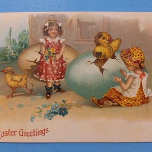 May include: An antique Easter postcard featuring two girls in dresses with polka dots. One girl is holding a bouquet of flowers. The other girl is sitting next to a large blue egg with a yellow chick inside. The text "Easter Greetings" is printed at the bottom of the card.