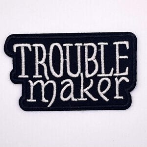 May include: A black embroidered patch with the words "TROUBLE maker" in white, stylized lettering. The patch has a black border and is rectangular with rounded corners. The text is centered on the patch.
