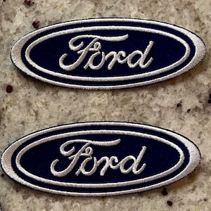 May include: Two oval-shaped embroidered patches featuring the Ford logo. Each patch has a dark navy blue background with the word Ford stitched in white script lettering, surrounded by a white embroidered border in an oval shape.
