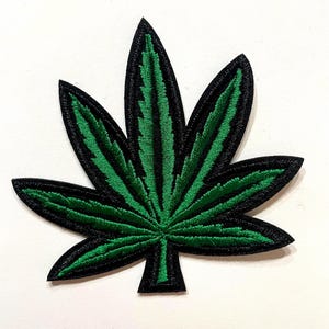 May include: Embroidered patch in the shape of a cannabis leaf. The leaf is green with black detailing and a black border. The patch is likely for clothing or accessories.