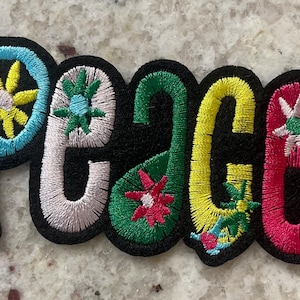 May include: Embroidered patch with the word "PEACE" in colorful, stylized letters. The letters are outlined in black and filled with various colors, including blue, white, green, yellow, and red, with floral accents.