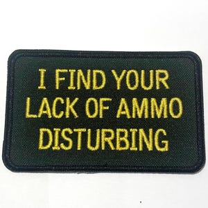 May include: A rectangular patch with a dark green background and a black border. The text "I FIND YOUR LACK OF AMMO DISTURBING" is embroidered in gold thread across the center of the patch.