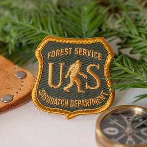 May include: A shield-shaped embroidered patch with a dark green background and gold border. The text reads FOREST SERVICE at the top and SASQUATCH DEPARTMENT at the bottom, with the letters U and S flanking a gold silhouette of a Sasquatch figure.