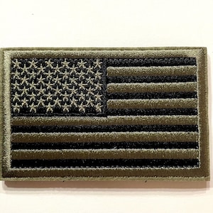 May include: An olive green and black embroidered patch depicting the American flag. The rectangular patch shows the stars and stripes in detail. The stars are in a black field, and the stripes alternate between olive green and black.