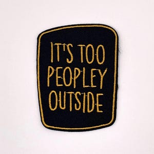 May include: Black patch with gold embroidered text that reads "IT'S TOO PEOPLEY OUTSIDE." The patch has a gold border and a rectangular shape with rounded corners. The patch is likely for clothing or accessories.