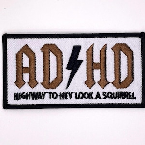 May include: Embroidered patch featuring the text "AD/HD" in brown gothic lettering, a black lightning bolt, and the phrase "HIGHWAY TO HEY LOOK A SQUIRREL" in black. The patch has a white background and a black border.