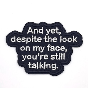 May include: A navy blue embroidered patch with white text. The text reads "And yet, despite the look on my face, you're still talking."