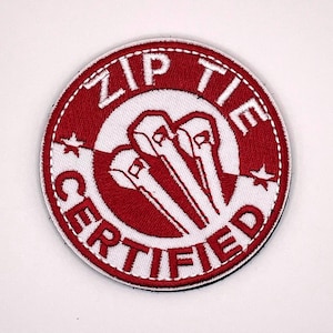 May include: A circular embroidered patch featuring a red and white design. The words "ZIP TIE CERTIFIED" are in red, encircling a white graphic of zip ties. The design also includes small stars.