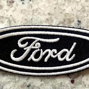 May include: An oval-shaped, black and white embroidered Ford patch. The patch features the word "Ford" in white script lettering, framed by a white border on a black background. Ideal for clothing or accessories.