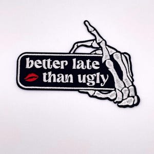 May include: Embroidered patch featuring a black rectangle with the phrase "better late than ugly" in white, a red kiss mark, and a white skeleton hand holding it. The patch is designed for clothing or accessories.