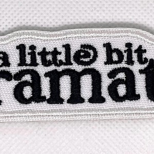 May include: A white embroidered patch featuring the text "a little bit dramatic" in black. The patch has a white, cloud-shaped border. Ideal for attaching to clothing or accessories.