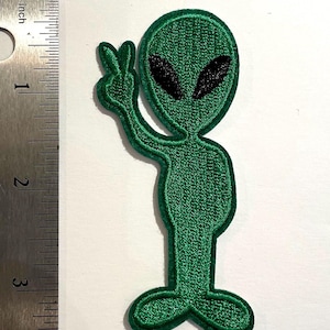 May include: A green embroidered alien patch featuring a peace sign gesture with one hand and large black oval eyes. The patch is positioned next to a metal ruler, showing the figure stands approximately 3.5 inches in total height.
