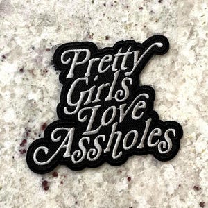 May include: Black embroidered patch featuring the phrase "Pretty Girls Love Assholes" in white cursive lettering. The patch has a black outline and is designed for application to fabric items, such as clothing or bags.