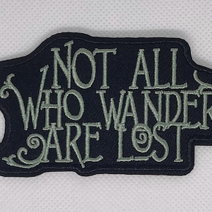 May include: A dark blue patch with the embroidered phrase "NOT ALL WHO WANDER ARE LOST" in light green. The patch has a unique shape with rounded edges and decorative swirls.