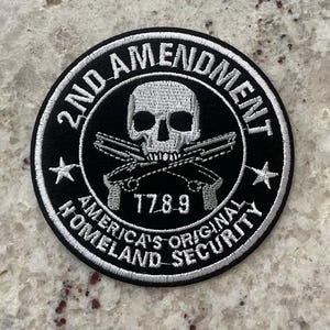 May include: A black and white embroidered patch with a skull and crossbones design. The text "2nd Amendment" is written in a circle around the skull and crossbones. The text "America's Original Homeland Security" is written in a circle around the outer edge of the patch. The year "1789" is written below the skull and crossbones.