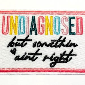 May include: Embroidered patch with the words "UNDIAGNOSED" in colourful letters above the phrase "but somethin' ain't right" in black script. The patch has a white background with a pink border.