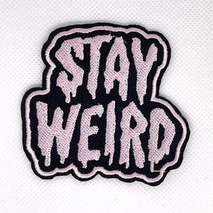May include: Embroidered patch with the words "STAY WEIRD" in light pink, dripping font on a black background. The patch has a black border and is likely for clothing or accessories.