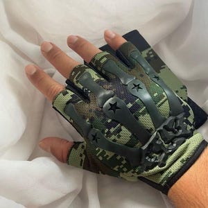 May include: Camouflage fingerless gloves with black accents and star details. The gloves feature a green, brown, and black digital camouflage pattern. The gloves are designed for protection and grip.