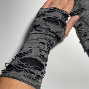 May include: Gray fingerless gloves with distressed, ripped detailing. The gloves are made of a soft, knit material and feature open areas throughout the design. The gloves cover the hand and extend up the forearm.