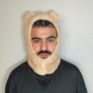 Peut inclure: Une personne portant un couvre-chef en polaire beige en forme de tête d'ours avec des oreilles. Le couvre-chef est porté sur la tête et le cou, et a une texture duveteuse.
