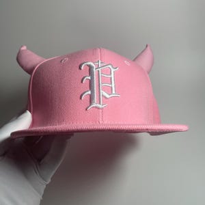 May include: Pink baseball cap with white devil horns and a white embroidered letter "P" on the front.
