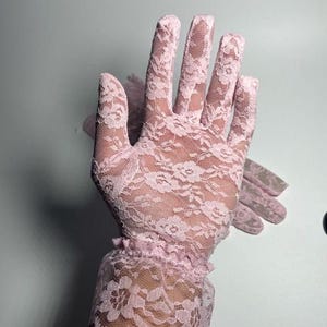May include: A pair of pink lace gloves with a ruffled cuff. The gloves are made of delicate lace with a floral pattern.