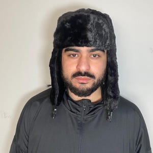 May include: A black faux fur trapper hat with ear flaps and a zip closure. The hat is being worn by a person with a beard.