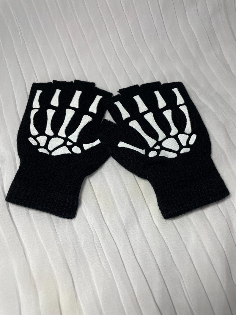 May include: A pair of black fingerless gloves with a white skeleton design. The gloves are made of a soft, knitted fabric.