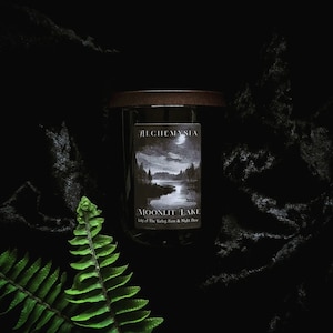 May include: A glass candle jar with a wooden lid, displaying a label with the text "ALCHEMYSIA" and "MOONLIT LAKE". The label shows a grayscale landscape of a lake and trees. A green fern leaf is in the foreground against a black backdrop.