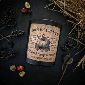 May include: Black candle with a label reading "Jack O' Lantern" and "Buttered Pumpkin Scone." The label features a pumpkin illustration. The candle is surrounded by decorative elements on a dark surface.