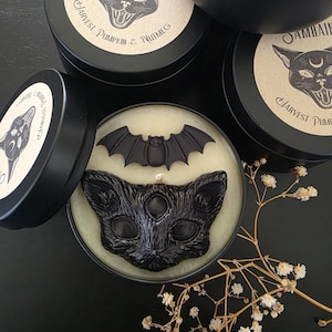 May include: A round black metal tin containing a candle with a black wax cat head and a black wax bat. The lid features a beige label with a black cat illustration and the text Samhain Harvest Pumpkin & Nutmeg. Additional tins are visible nearby.