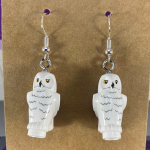 May include: A pair of white owl earrings with silver hooks. The owls have yellow eyes and gray feathers.