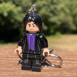 May include: Black Lego minifigure keychain with a black jacket, purple shirt, and black pants. The minifigure has black hair and a serious expression.